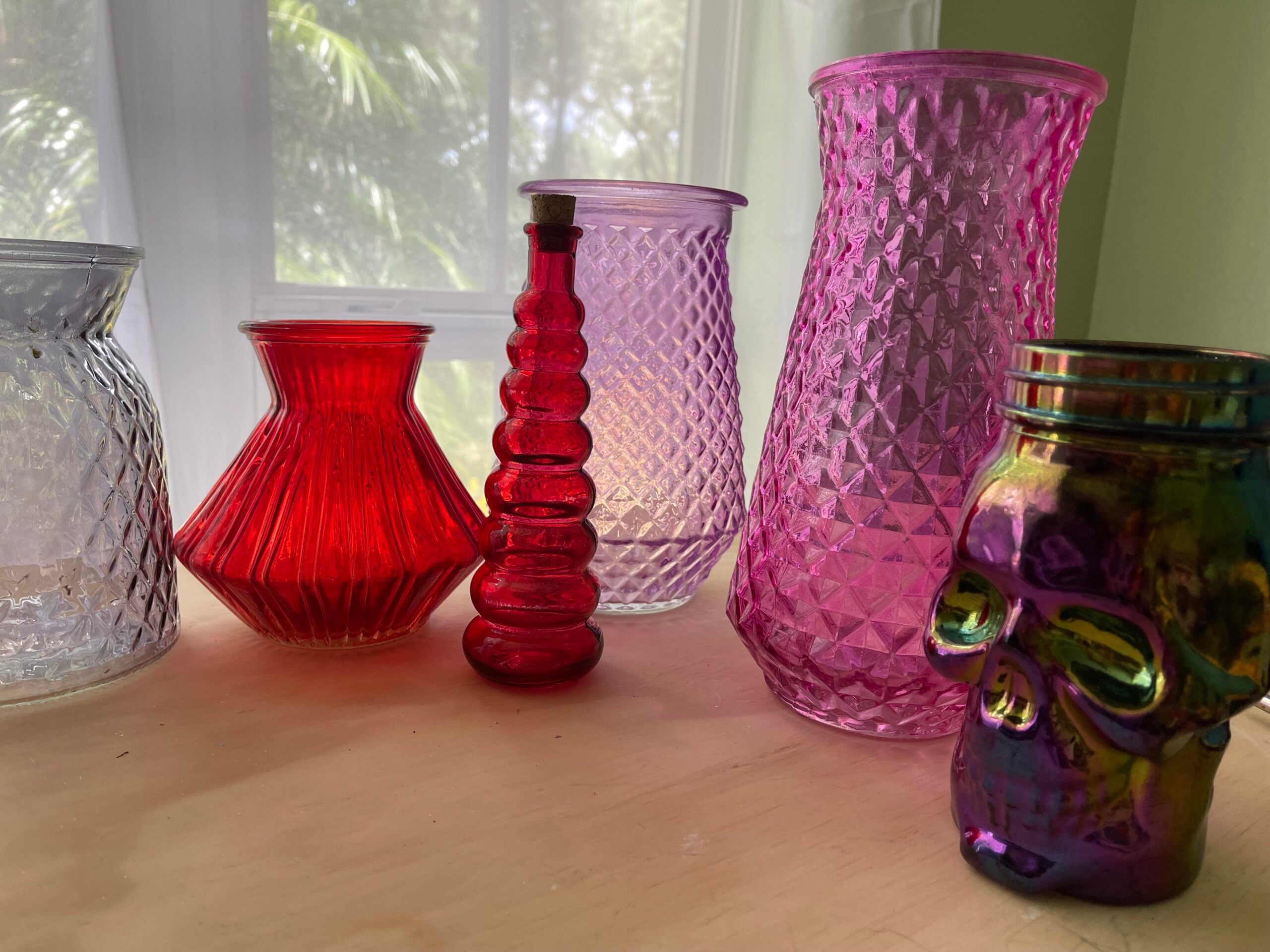 A group of colorful glass vases and jars, including red, purple, clear, and an iridescent skull-shaped jar, are arranged on a wooden surface in sunlight near a window with white curtains.