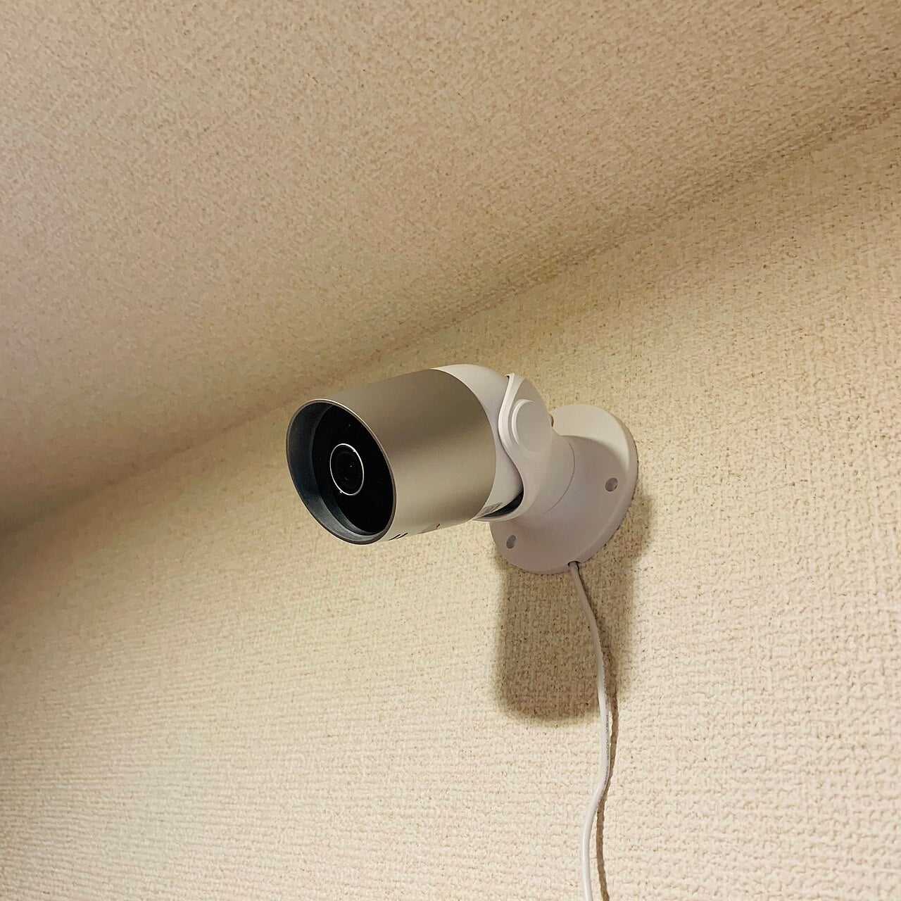A white security camera is mounted on a textured beige wall near the ceiling, with its power cable hanging down.