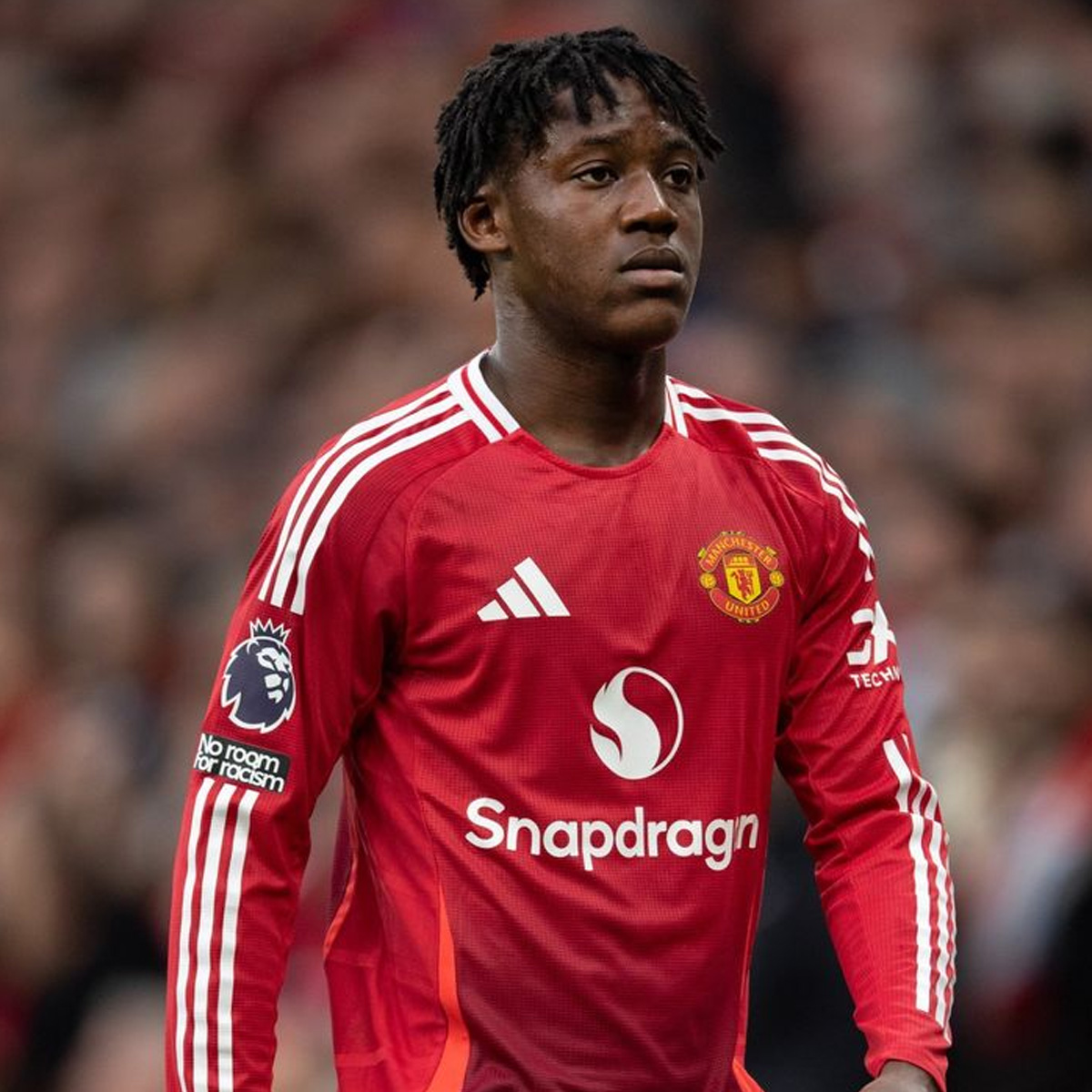 A football player wearing a red Manchester United jersey with "Snapdragon" sponsor and "Premier League" patch stands on the field, looking ahead with a focused expression.