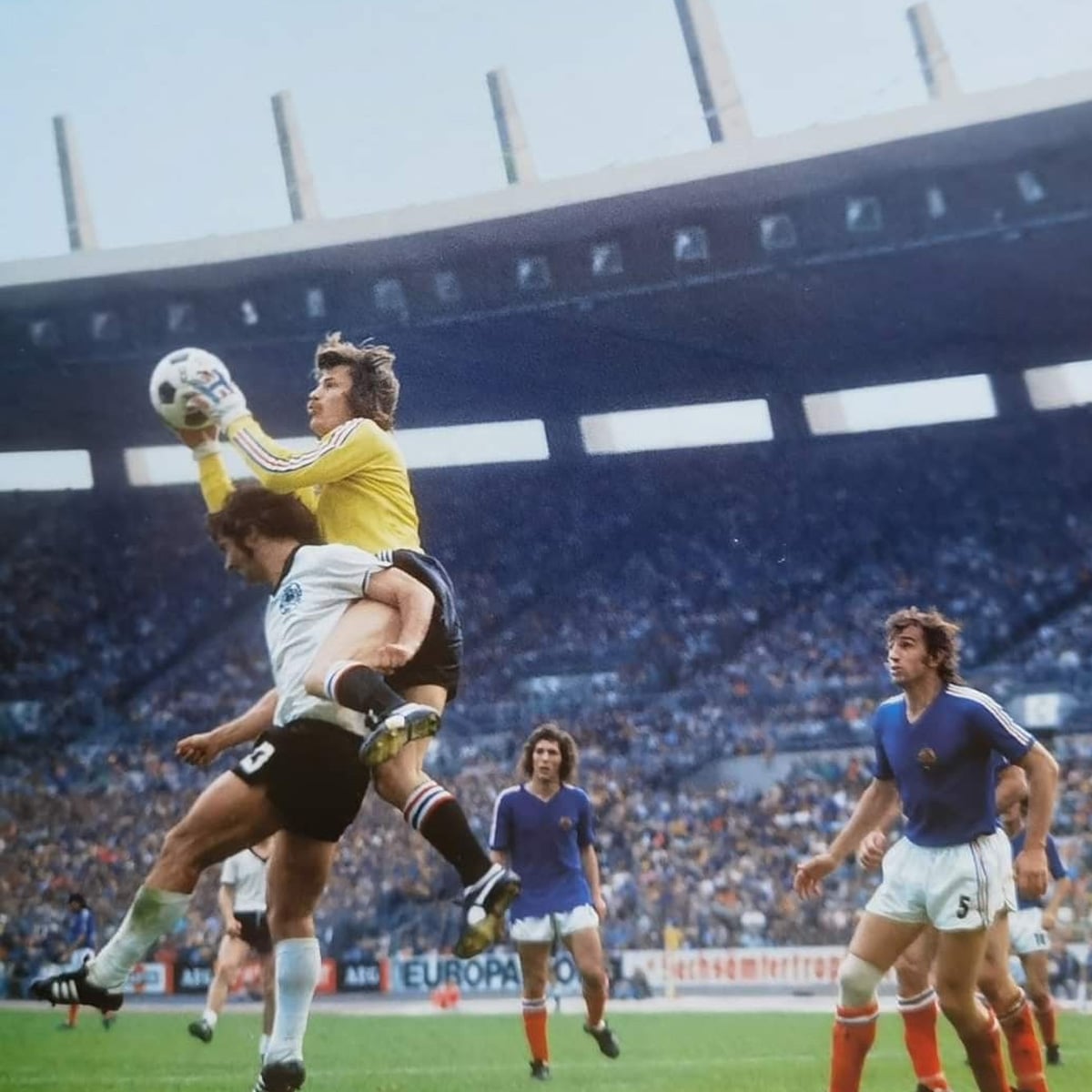 A soccer goalkeeper in yellow jumps to catch the ball, colliding with an opposing player in white and black. Other players in blue and white watch, with a large crowd in the stadium stands in the background.