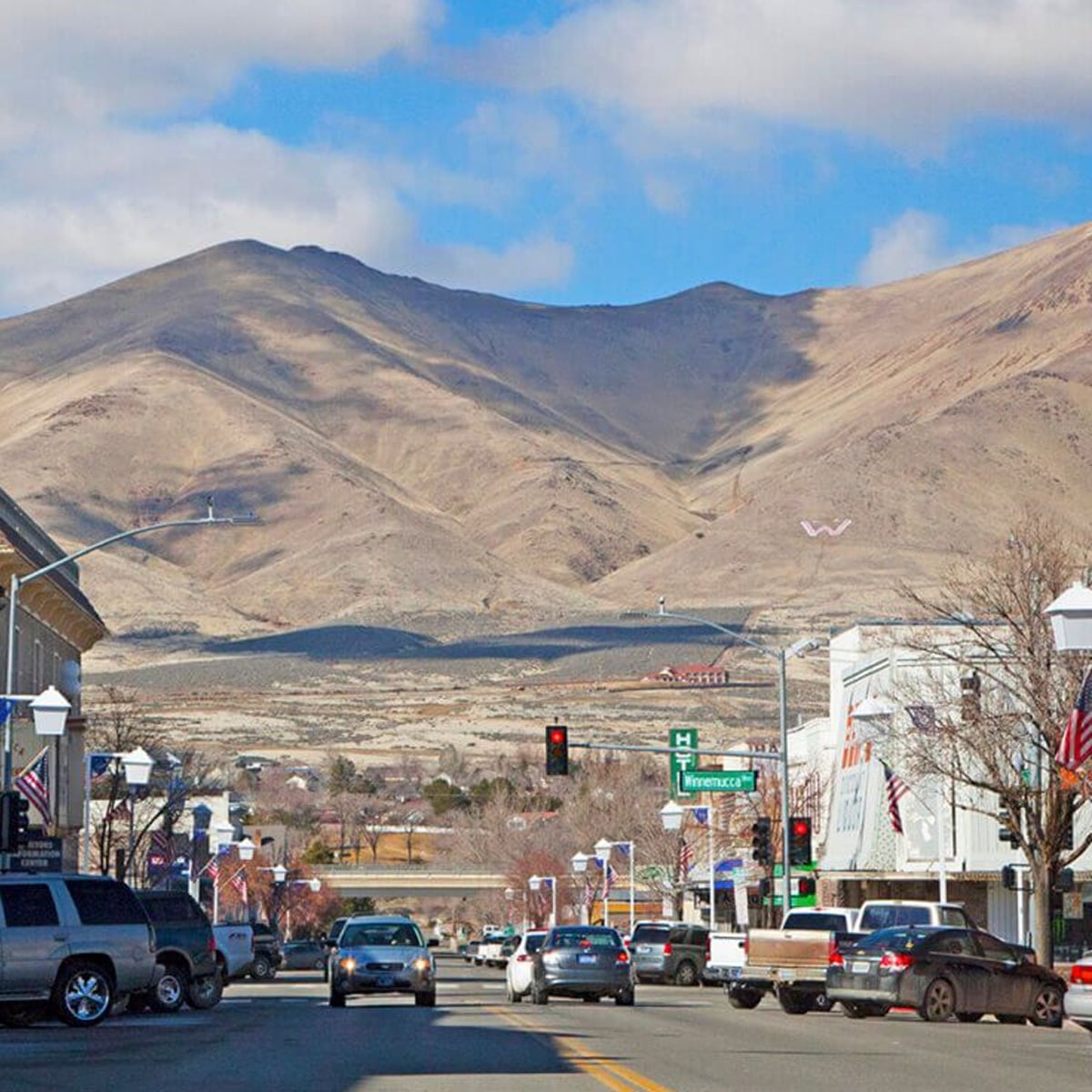 A small town street lined with cars and buildings leads toward dry, brown mountains under a partly cloudy sky. American flags hang from lampposts, and a large white "W" is visible on the hillside in the distance.