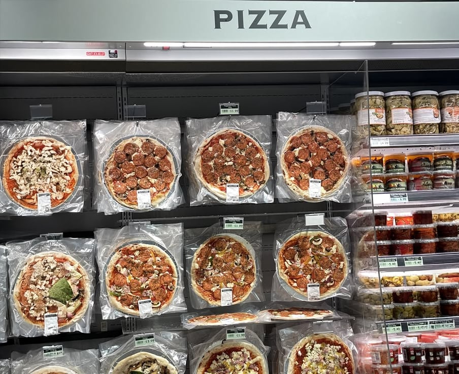 A grocery store shelf displays various packaged pizzas in clear plastic wrappers, arranged in rows. The section is labeled "PIZZA" above, and jars of condiments are visible on the right side.