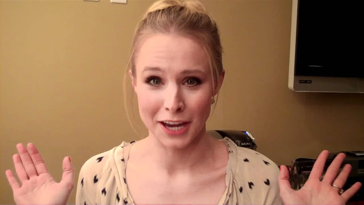A woman with blonde hair in a bun smiles and raises both hands beside her face, palms forward. She is indoors, wearing a light-colored top with a small pattern, and stands against a beige wall.