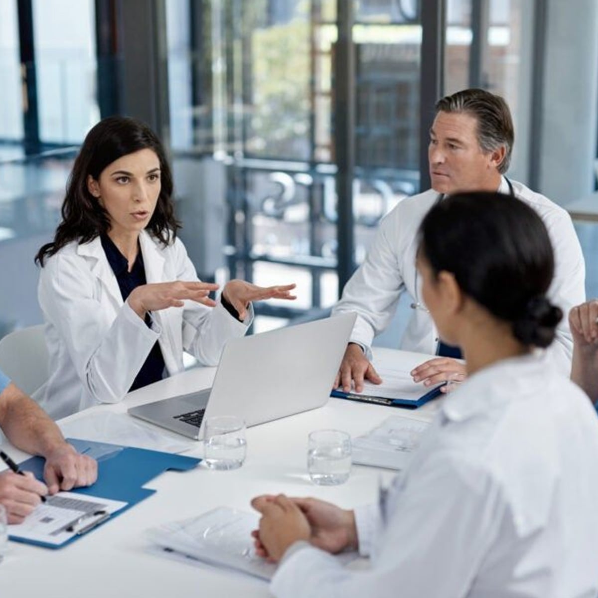 A group of doctors in white coats sits around a table in a modern office, engaged in discussion. One woman speaks while others listen, with a laptop and documents on the table.