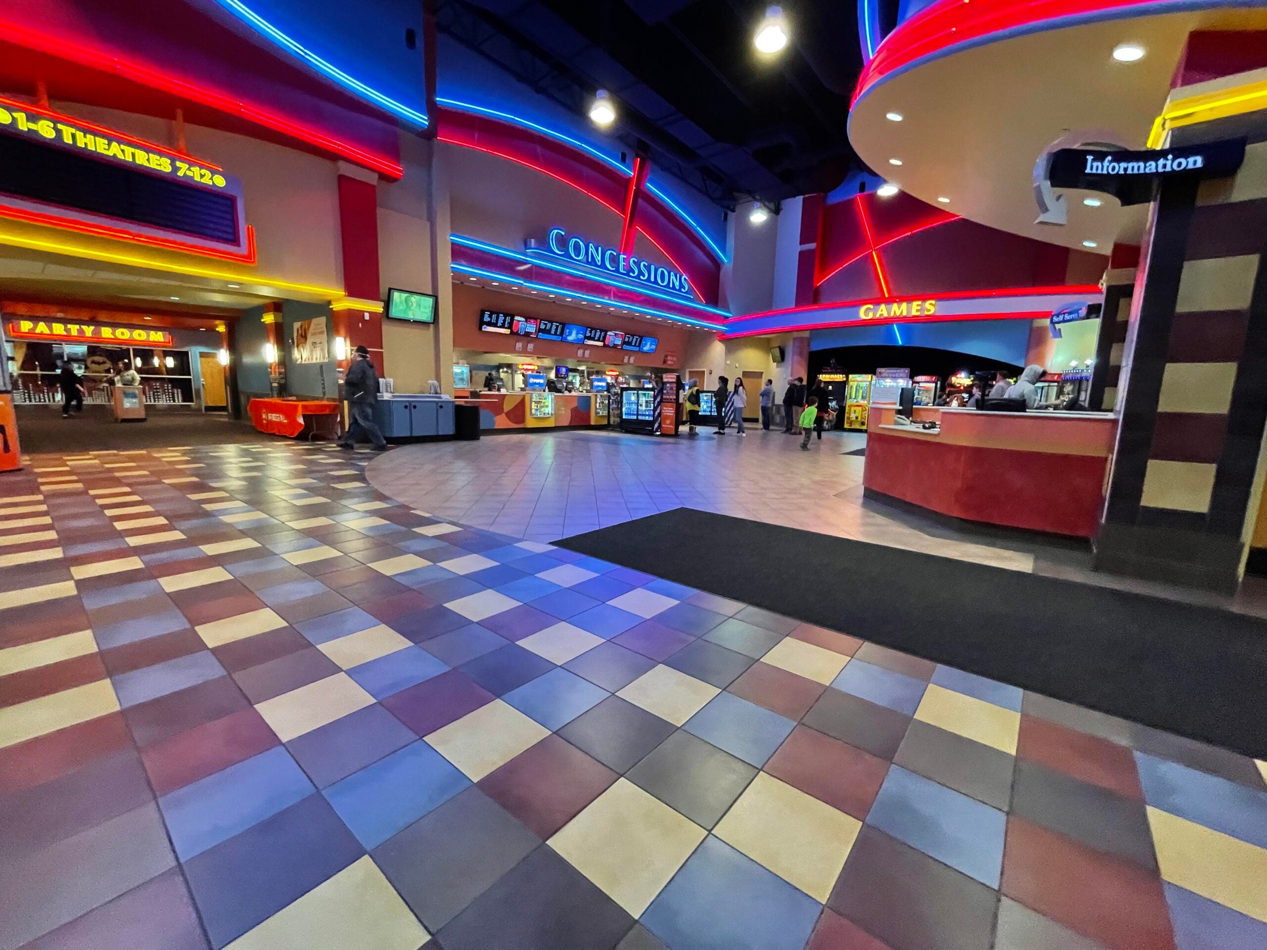 A brightly lit movie theater lobby with colorful neon lights, concession stands, a sign for games, tiled flooring, and movie listings above theater entrances. A few people are walking and standing around.