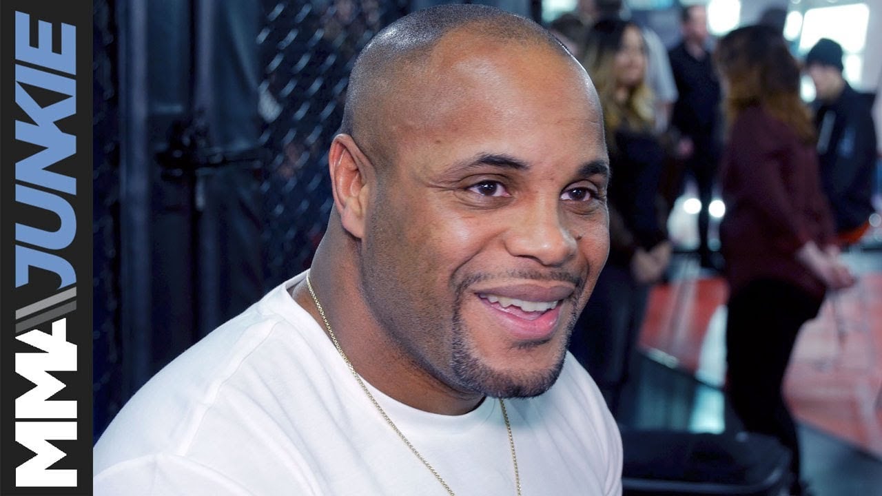 A man in a white shirt smiles during an interview at an MMA gym, with people and training equipment visible in the blurred background. MMA Junkie branding is displayed on the left side of the image.