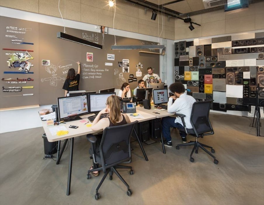 A modern office with six people working at desks with computers. The room has creative wall art, notes on a large chalkboard, and a wall decorated with speakers. The atmosphere appears collaborative and casual.