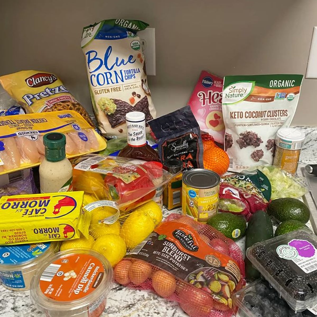 A kitchen counter displays various groceries, including fruits, vegetables, chicken, chips, keto coconut clusters, rice, salad dressing, pretzels, lemon, avocado, berries, canned goods, and other packaged foods.