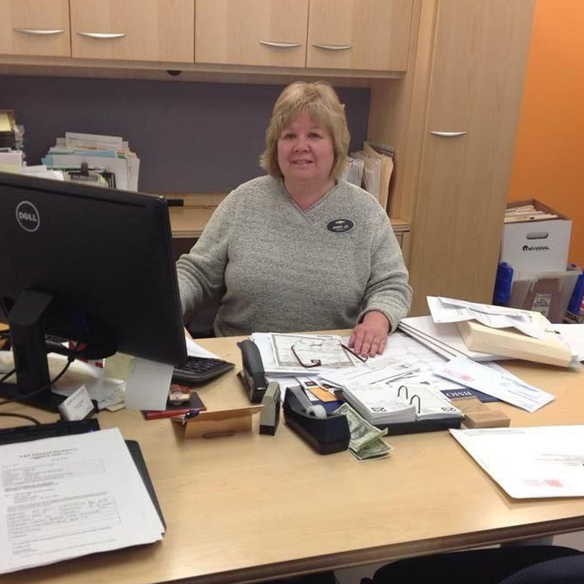 A woman with short blonde hair sits at a desk cluttered with papers, a computer monitor, cash, and office supplies. She is smiling and wearing a light gray sweater in an office with wooden cabinets and orange walls.