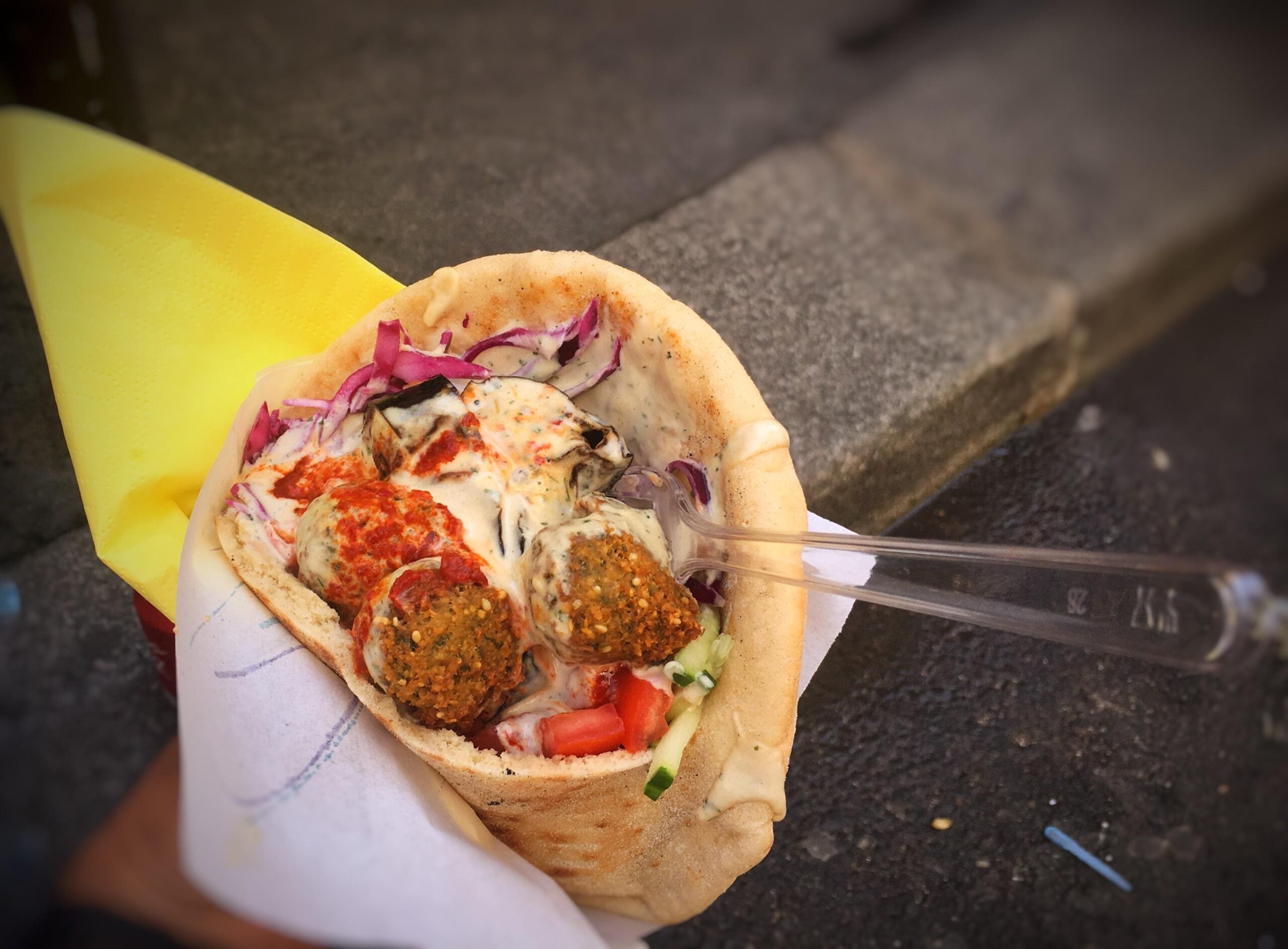 A pita filled with falafel balls, chopped tomatoes, cucumber, red cabbage, and creamy sauce, held in a paper wrap with a yellow napkin and a clear plastic fork, resting near a curb on a street.