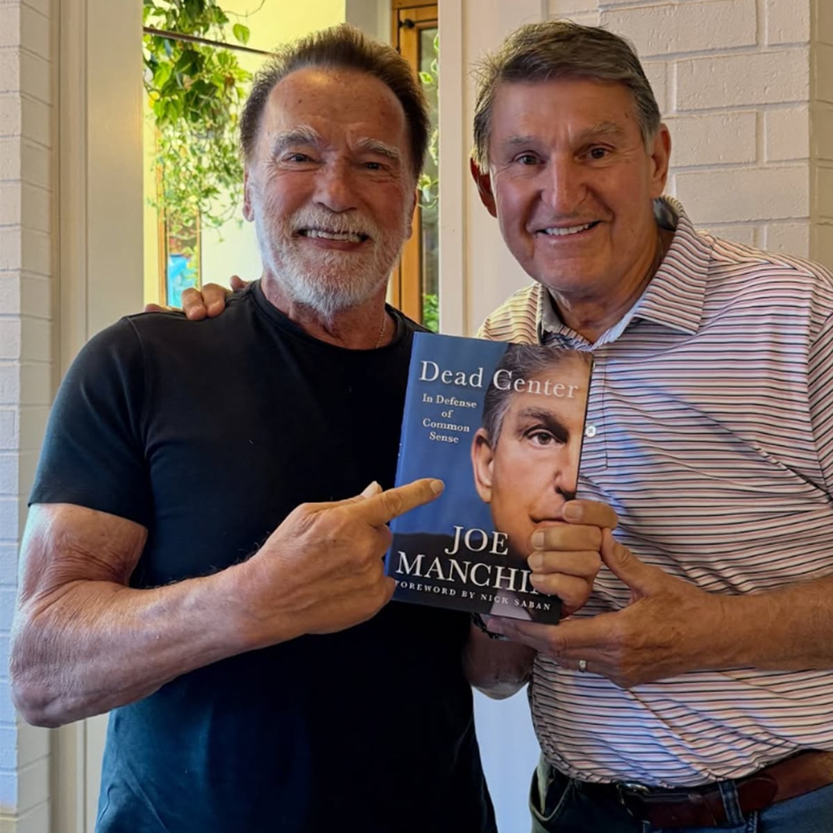 Two smiling men stand side by side. One man is holding and pointing to a book titled "Dead Center" by Joe Manchin. The background includes a white brick wall and greenery.