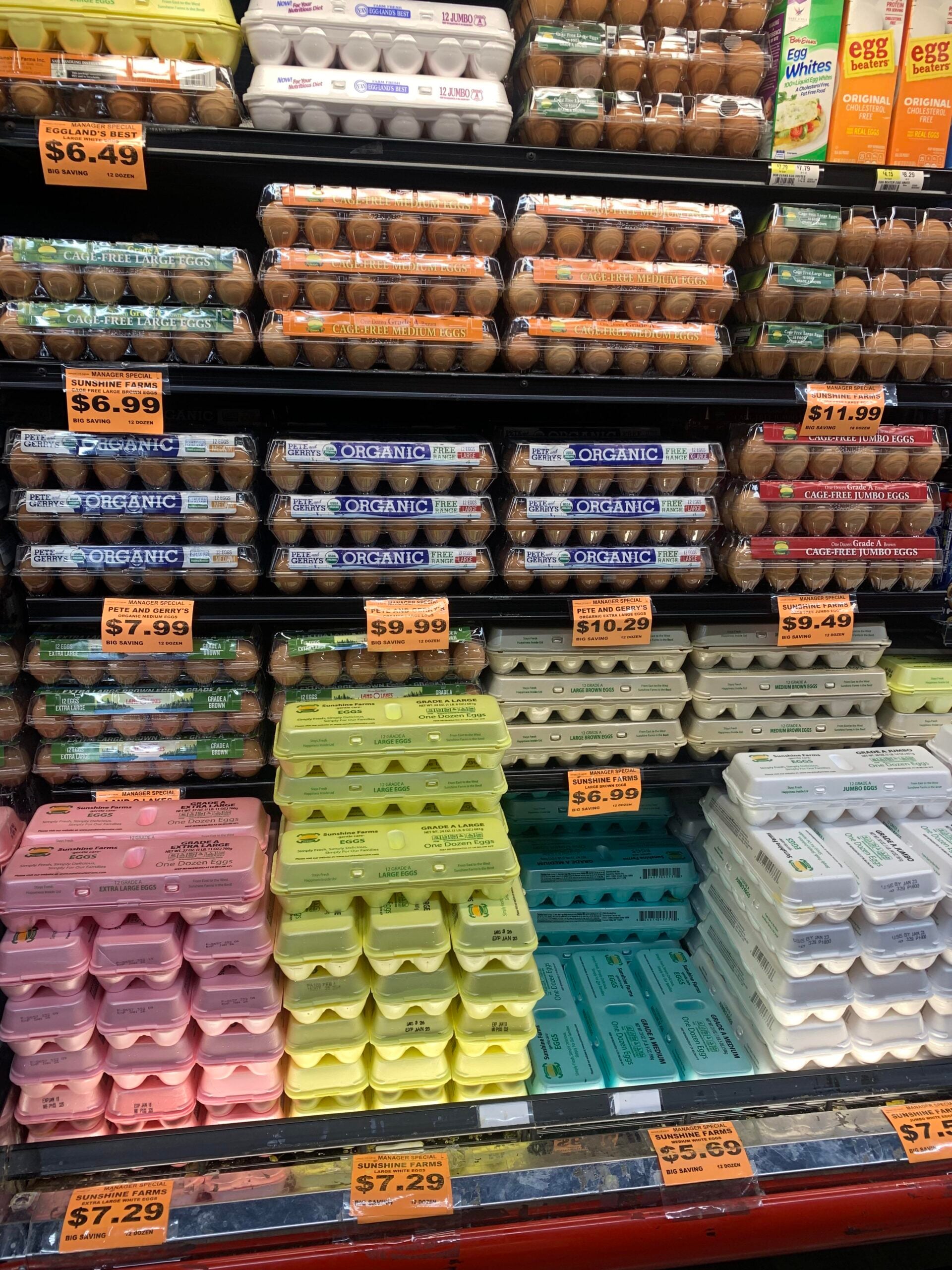 A grocery store refrigerator section filled with various brands and types of eggs in colorful cartons. Price tags are visible, with organic, free-range, and conventional options stacked in rows.