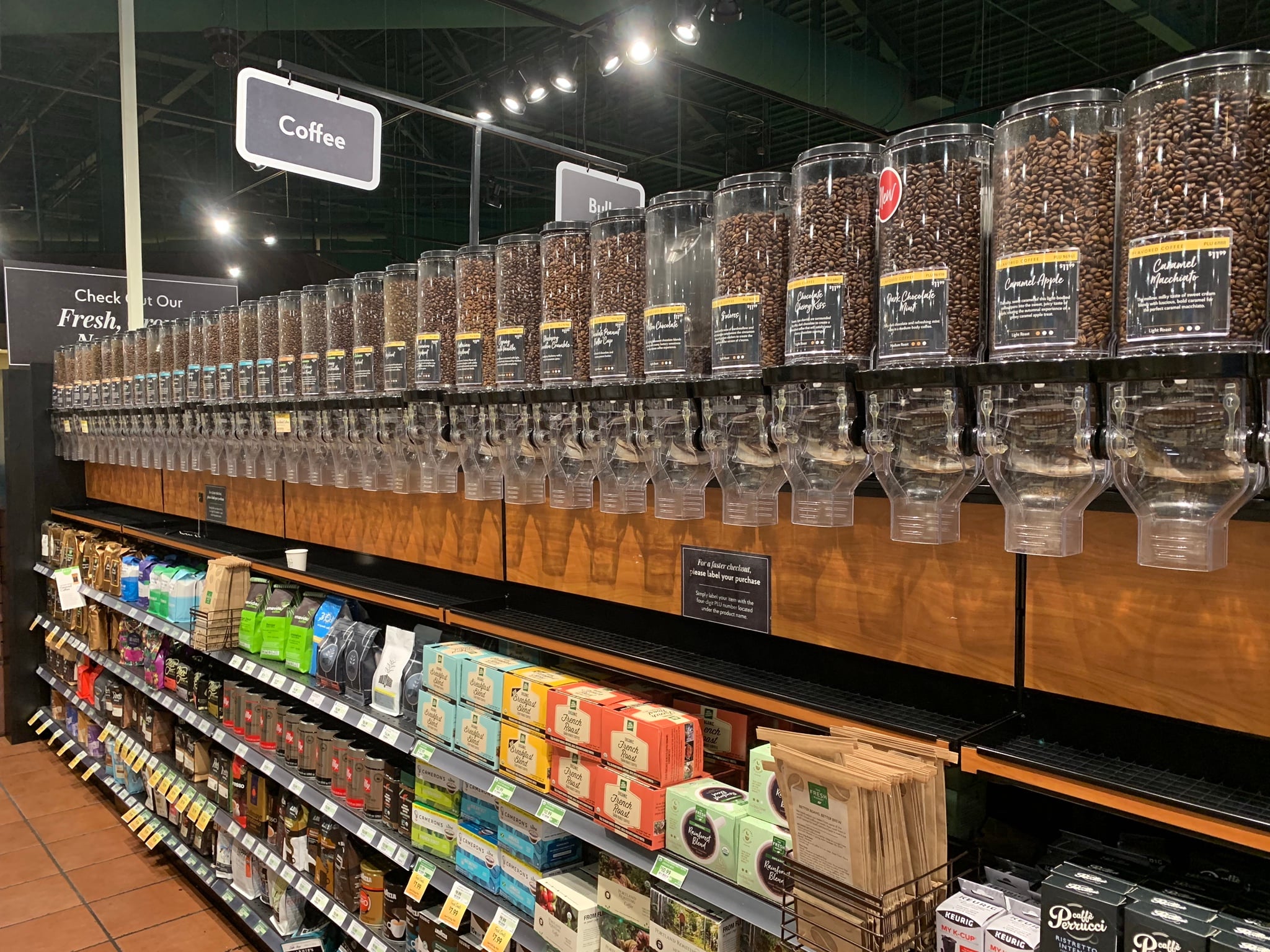 A grocery store aisle with rows of bulk coffee bean dispensers on the top shelf and various packaged coffee products and filters on shelves below. A hanging sign above reads “Coffee.”