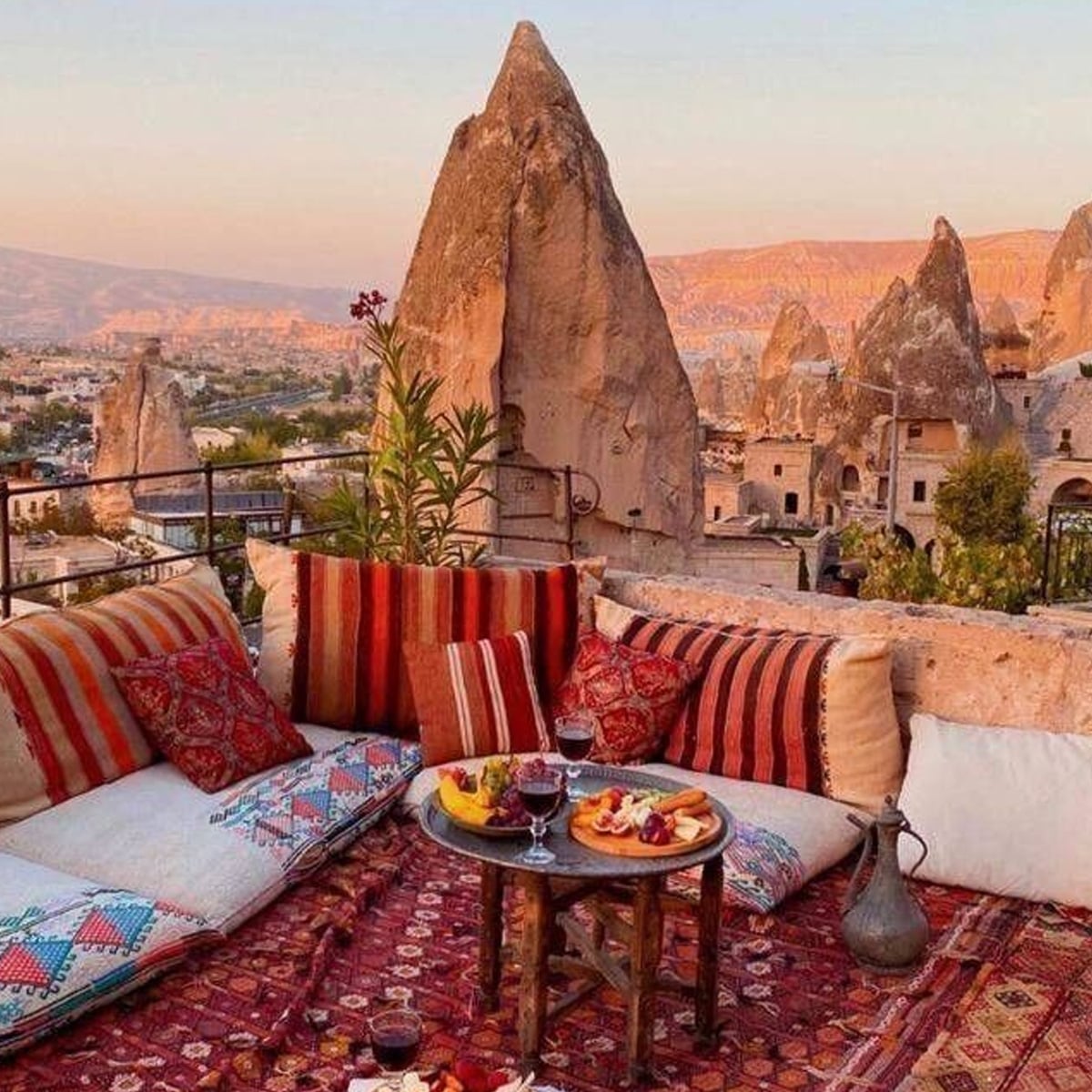 Cozy outdoor seating with colorful cushions and carpets, a table set with wine and fruit, overlooking the unique rock formations of Cappadocia, Turkey at sunset. Warm, scenic, and inviting atmosphere.