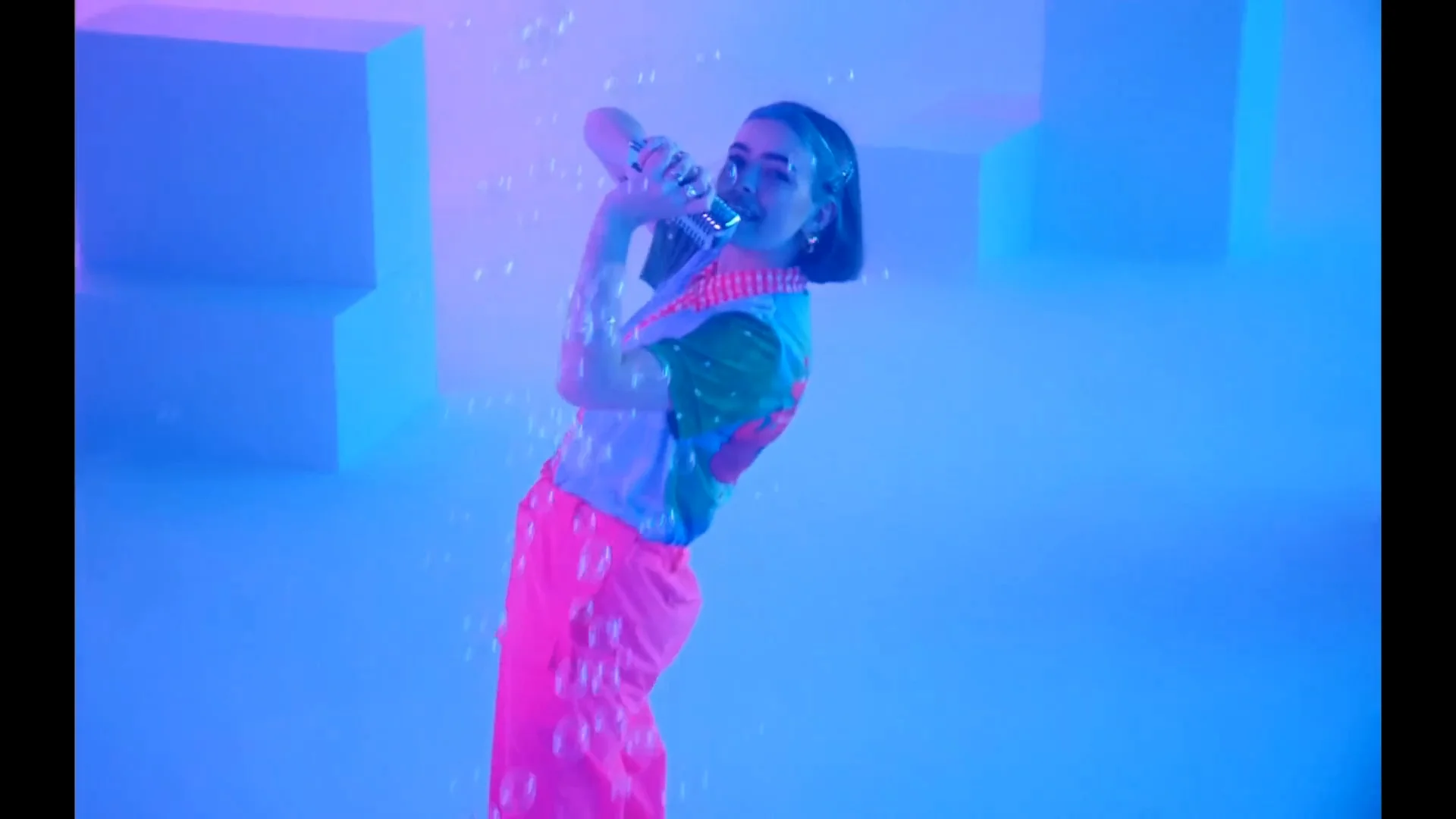 A woman in colorful clothing sings into a microphone, surrounded by floating bubbles, in a room lit with vibrant blue and pink lights and geometric shapes in the background.