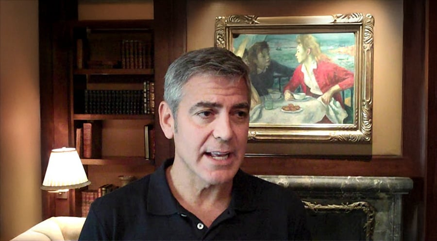 A man with short gray hair and a black shirt speaks in a warmly lit room, with bookshelves, a lamp, and a framed painting of two people dining on the wall behind him.