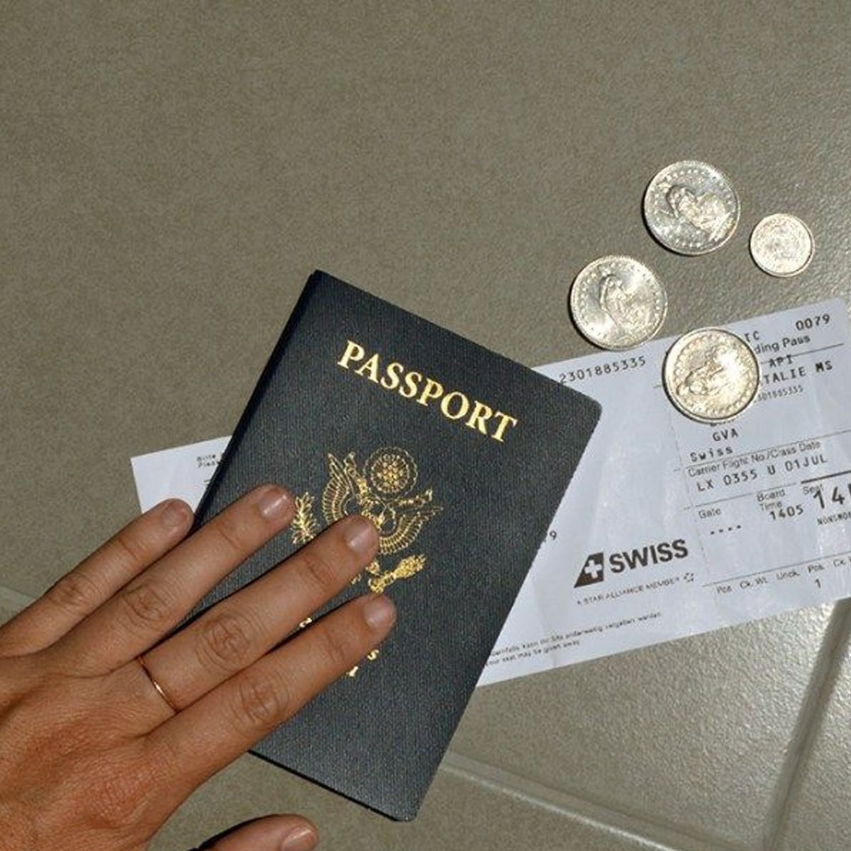A hand holding a U.S. passport rests on a boarding pass, with several coins scattered nearby on a table.