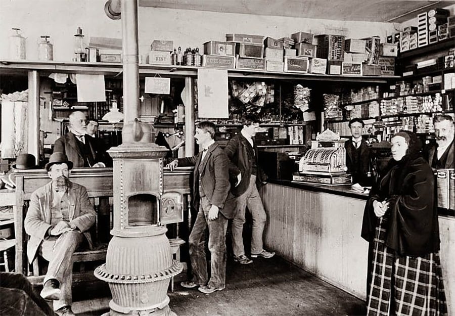 A black-and-white photo of a general store with men standing and sitting near a stove, shelves stocked with goods, a cash register on the counter, and a woman in plaid clothing near the front.