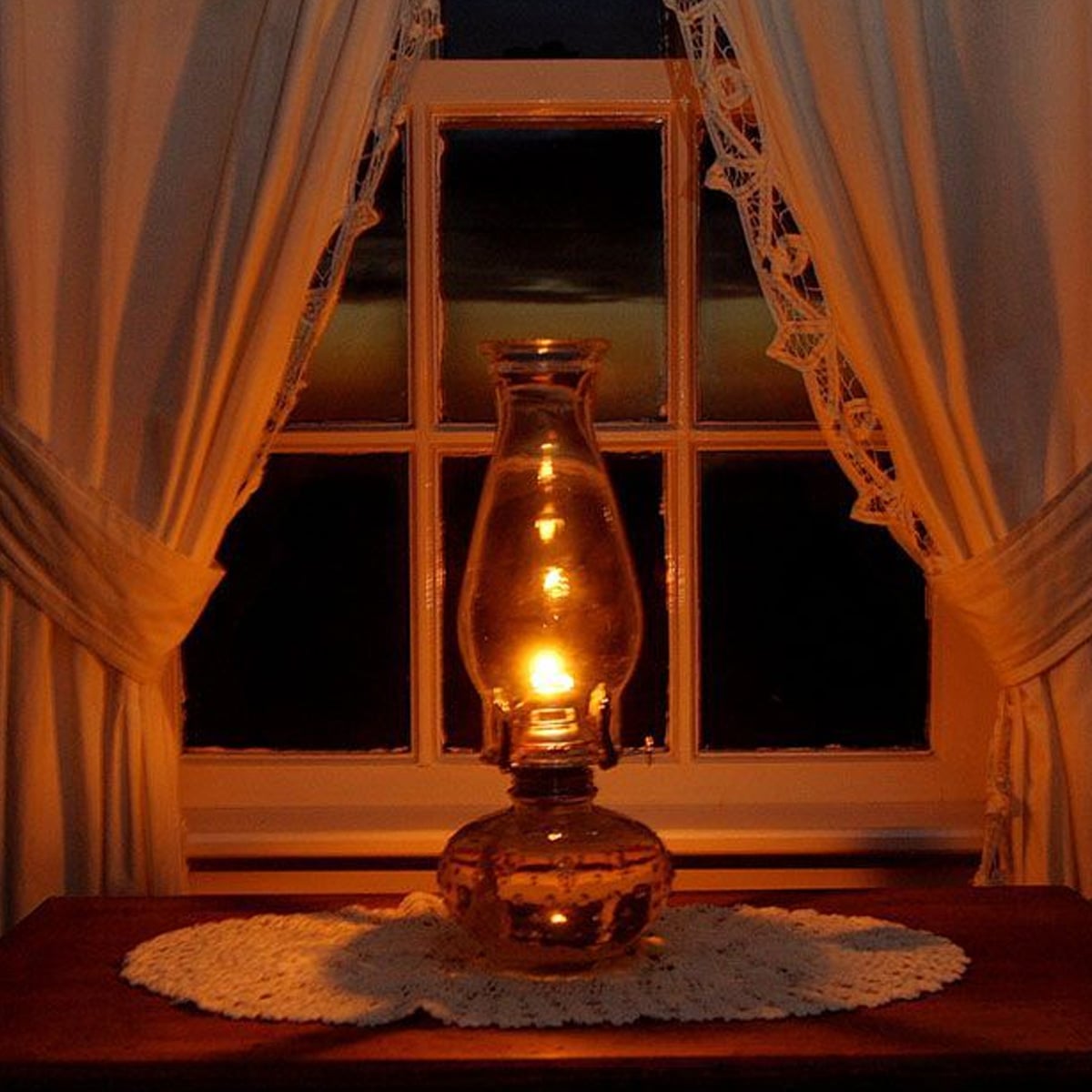 An oil lamp glows warmly on a lace doily in front of a window with white curtains, illuminating the dark night outside.
