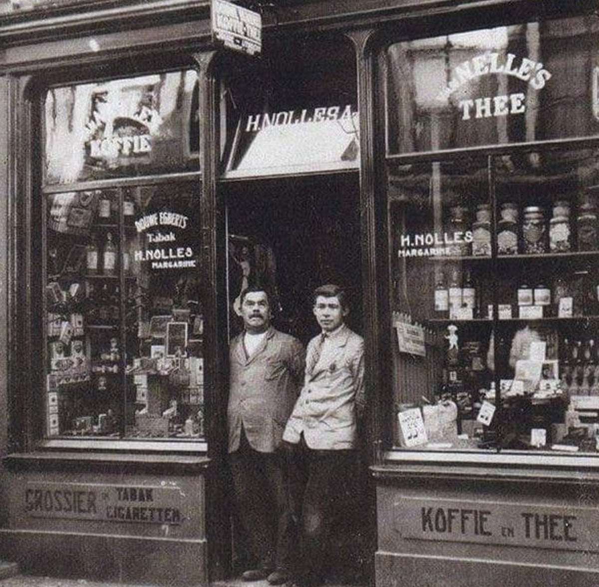 Two men stand in the doorway of an old-fashioned shop with large windows displaying various goods and signage, including "KOFFIE" and "THEE." The storefront is labeled "H. Nolles" and products fill the window displays.