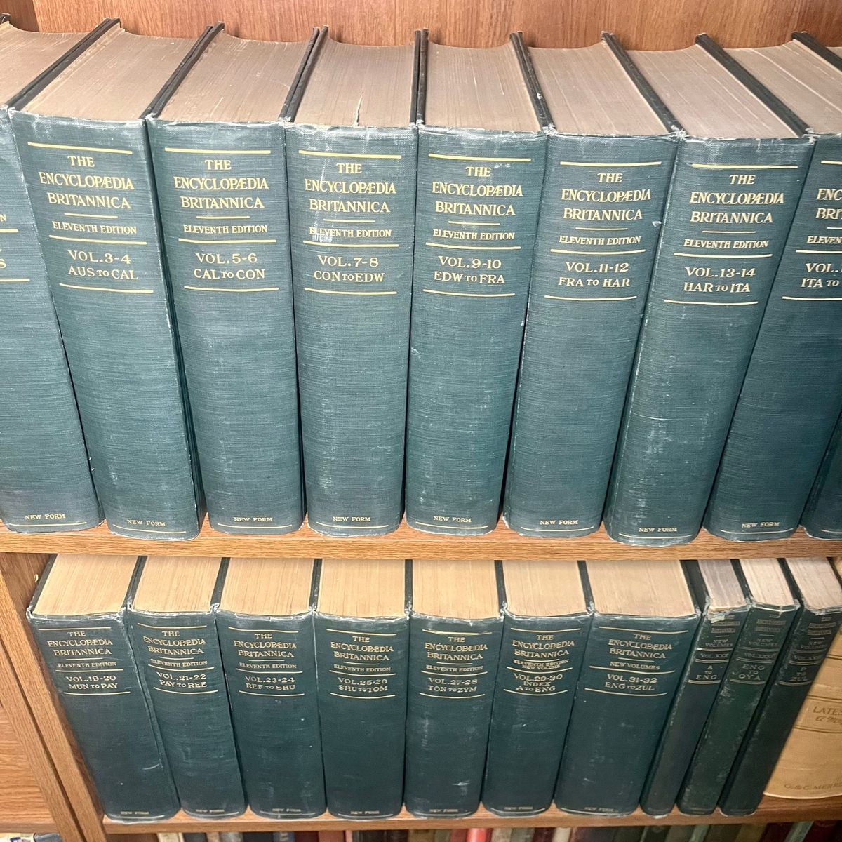 Several volumes of the Encyclopedia Britannica, Eleventh Edition, are neatly arranged on wooden shelves. The book spines are dark green with gold text, listing volume numbers and content ranges.