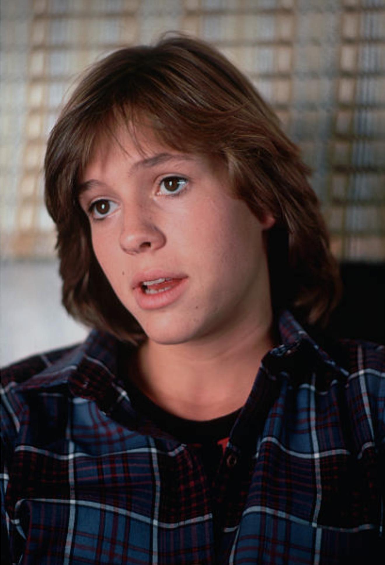 A young person with shoulder-length brown hair and a plaid shirt looks slightly to the side, mouth slightly open, against a blurred background with a woven texture.