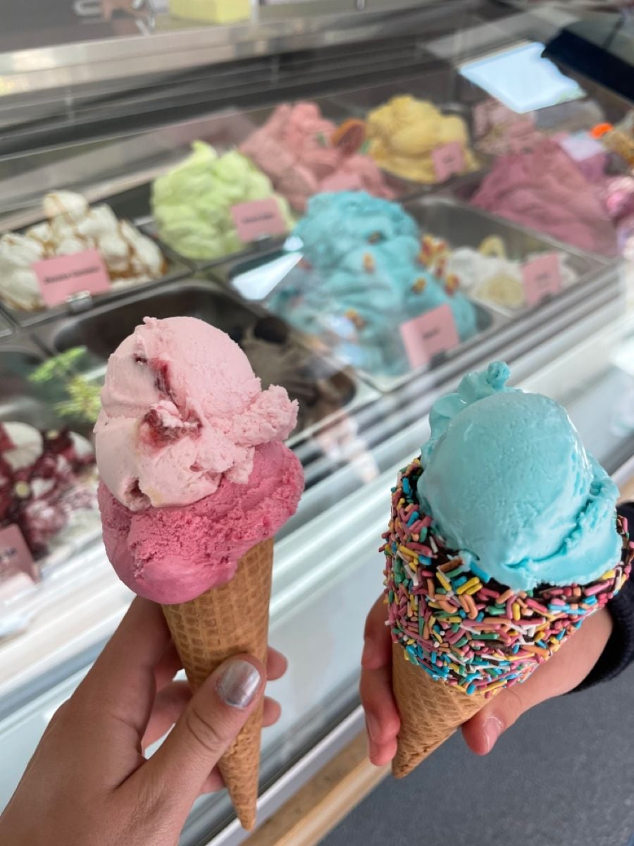 Two hands holding ice cream cones; one with pink ice cream, the other with blue ice cream and rainbow sprinkles. A display of various colorful ice cream flavors is visible in the background.