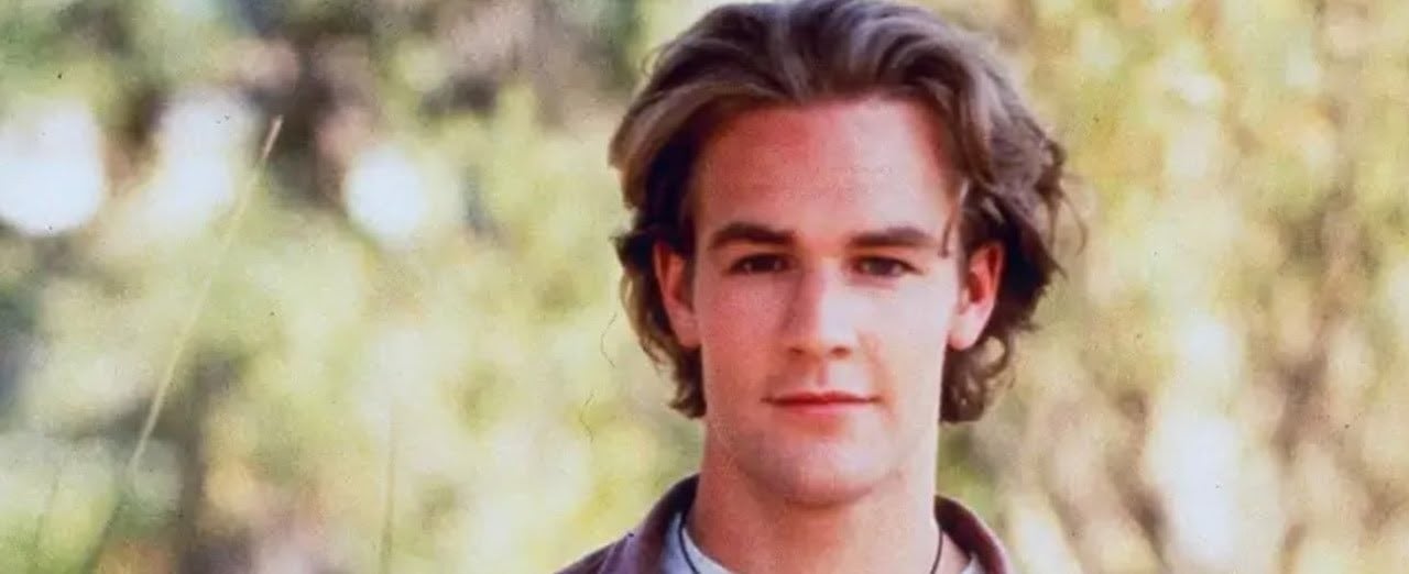 A young man with wavy brown hair and fair skin looks into the camera, standing outdoors with a blurred background of green and yellow foliage.