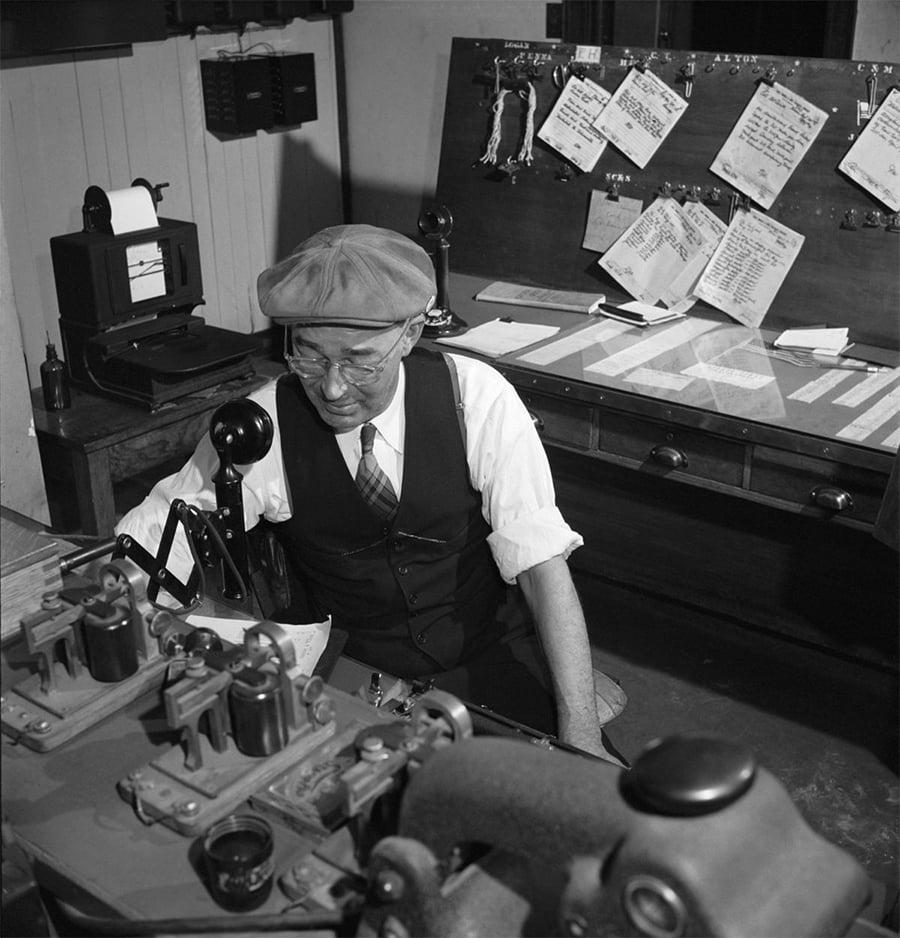 An older man wearing a vest, tie, and flat cap sits at a desk with vintage telecommunication equipment and papers. Behind him is a bulletin board with documents and a telephone on the desk. Black and white photo.