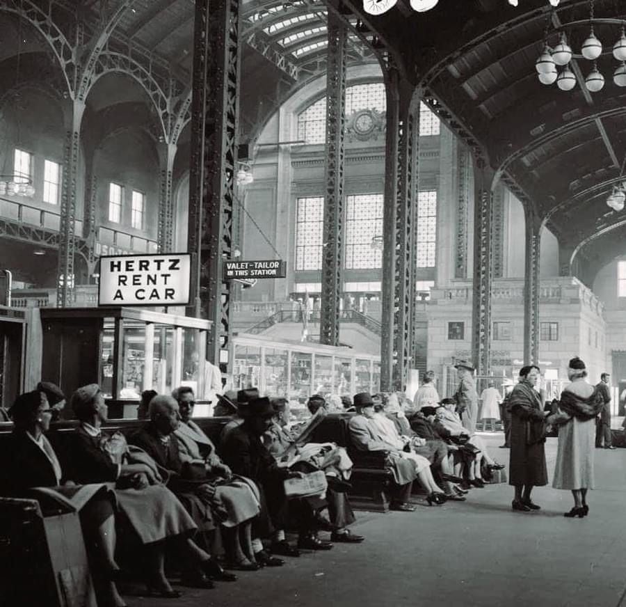 Black and white photo of a busy train station waiting area with people sitting and standing. A "Hertz Rent a Car" sign is visible. The architecture features tall arches and large windows.