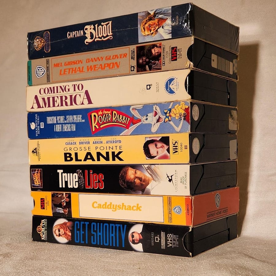 A stack of eight VHS tapes, including titles like Captain Blood, Lethal Weapon, Coming to America, Who Framed Roger Rabbit, Blank, True Lies, Caddyshack, and Get Shorty, displayed on a beige surface.