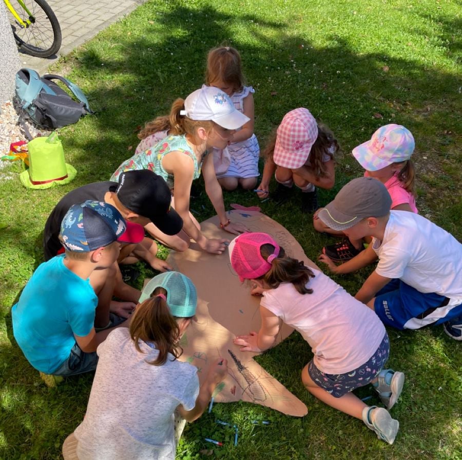 A group of young children wearing caps sit and kneel on grass outside, drawing and coloring on a large piece of brown paper shaped like a fish. Art supplies are scattered nearby. The scene is bright and sunny.
