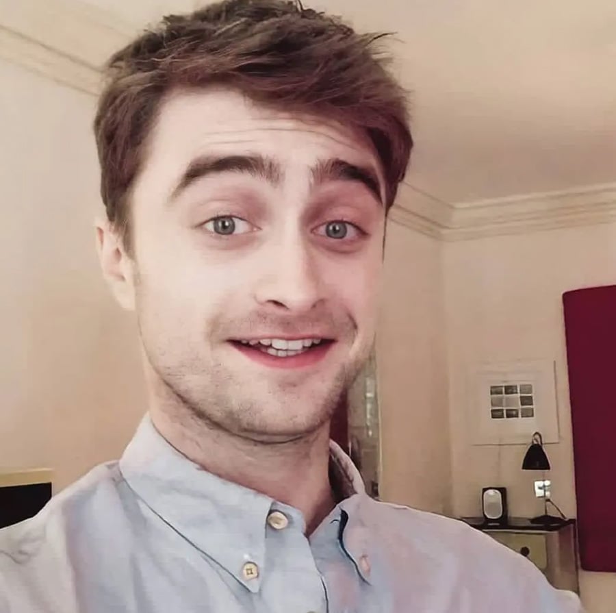 A young man with short brown hair and light eyes is smiling at the camera, wearing a light blue button-up shirt, standing in a softly lit room with cream-colored walls and minimal decor.