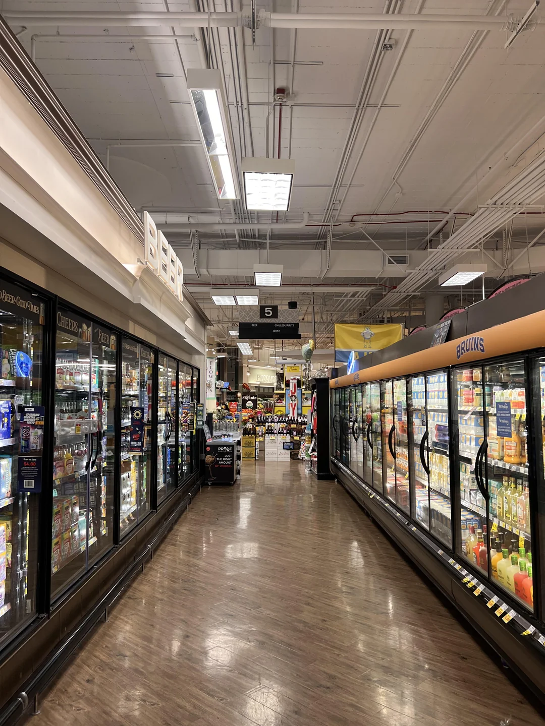 A grocery store aisle with refrigerated cases on both sides containing beer and dairy products, bright overhead lights, and shelves of various items visible at the end of the aisle.