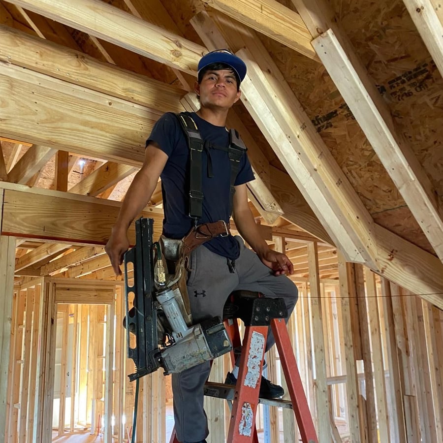 A construction worker wearing a blue cap, tool belt, and harness stands on a ladder inside a house frame with exposed wooden beams and studs.
