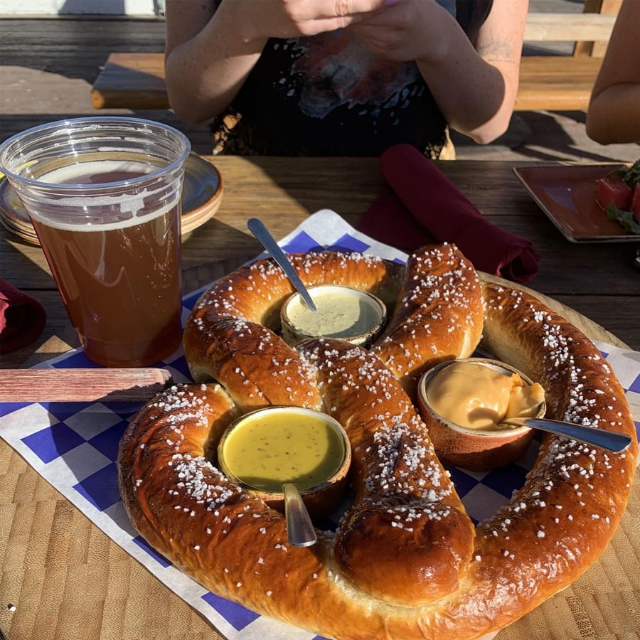 A large salted pretzel served on paper with two small bowls of dipping sauces and a glass of beer on a wooden table; a person sits in the background with hands over a plate.