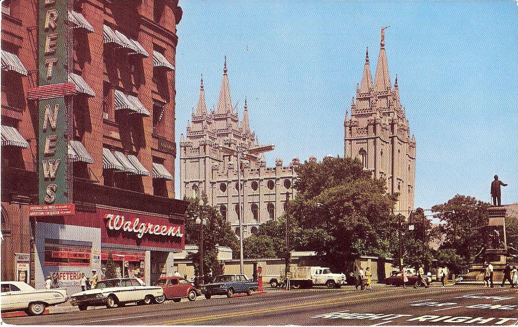 A city street scene with a Walgreens store and “Street News” sign on the left, cars parked along the road, and a large Gothic-style church with spires in the background. Trees and a statue add to the urban setting.
