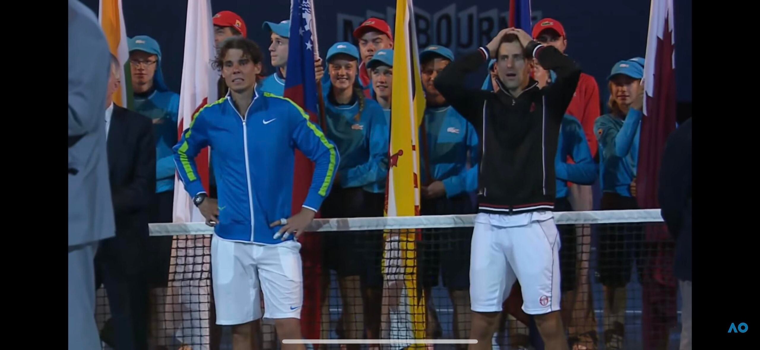 Two male tennis players stand at the net, one with hands on hips and the other with hands on his head, while ball kids and officials watch in the background during an outdoor tournament.