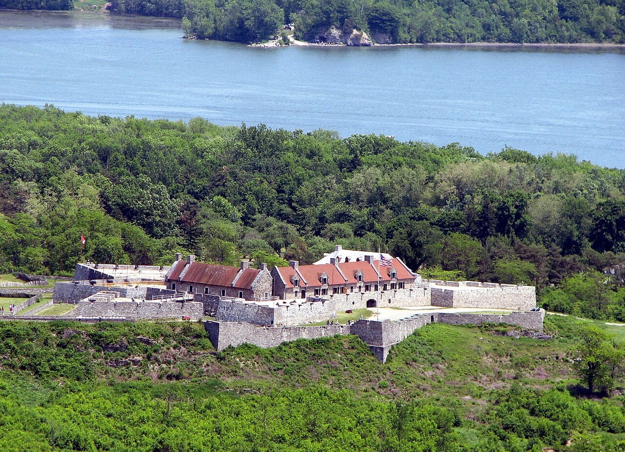 Aerial view of a historic stone fort with red-roofed buildings, surrounded by green trees, located near a river or large body of water.