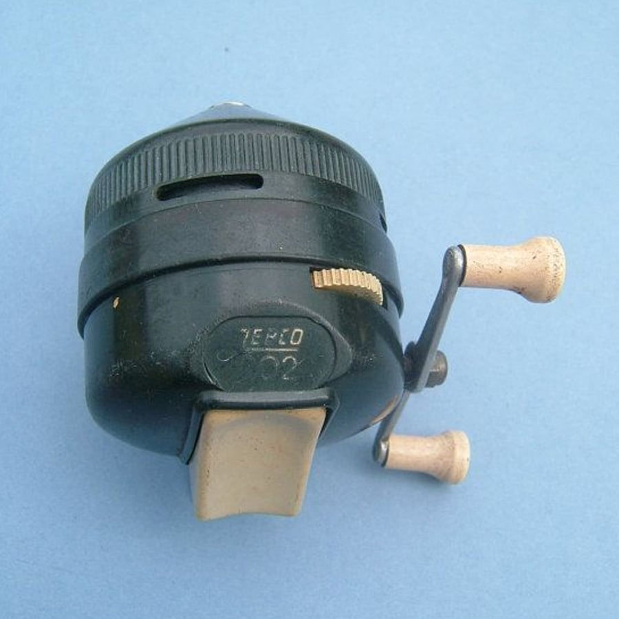 A vintage Zebco 202 fishing reel with a black body, cream-colored thumb button, and two beige handle knobs, set against a light blue background.