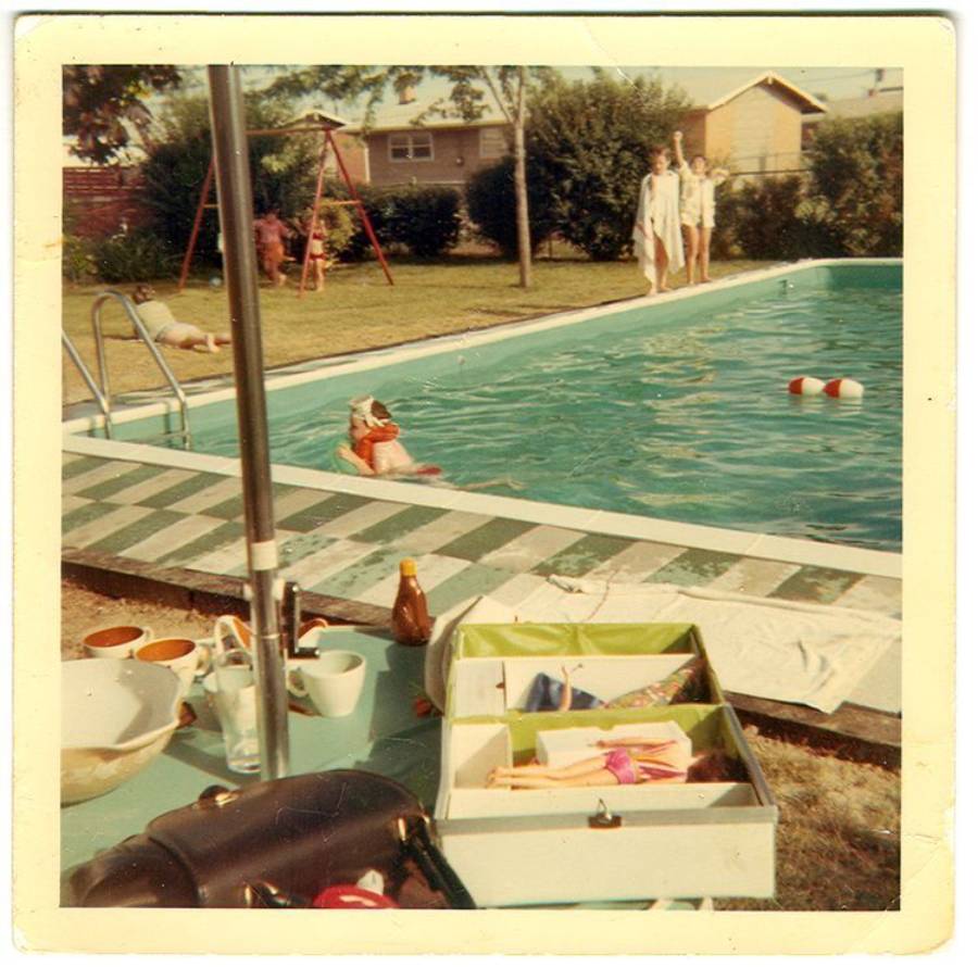 Old photograph of a backyard pool scene with people swimming and relaxing. In the foreground, a picnic setup with food and an open box is visible. In the background, kids play near a swing set and houses line the yard.