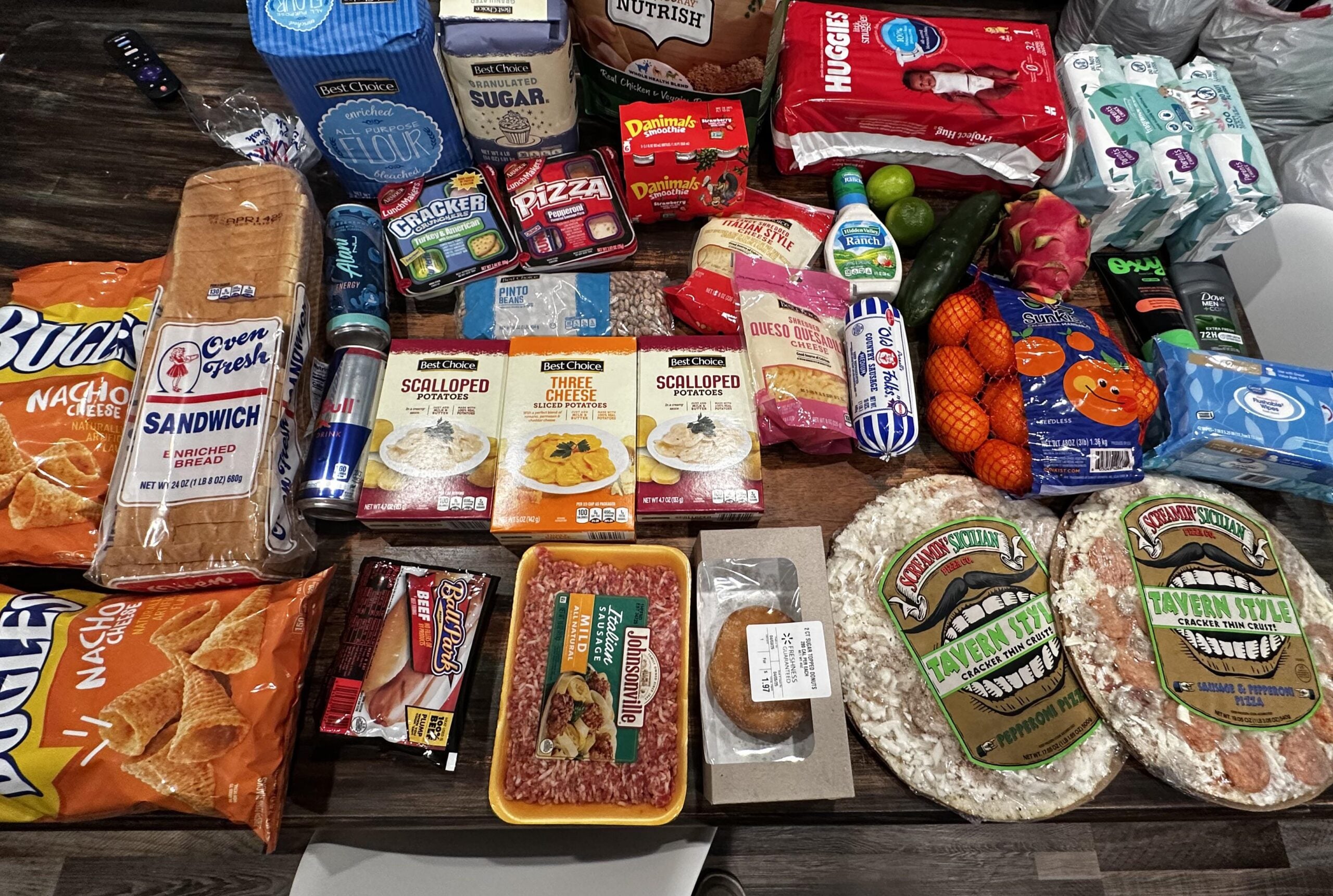A table is filled with assorted groceries, including bread, chips, pizzas, drinks, fresh produce, packaged meats, snacks, hygiene products like baby wipes, and household essentials.