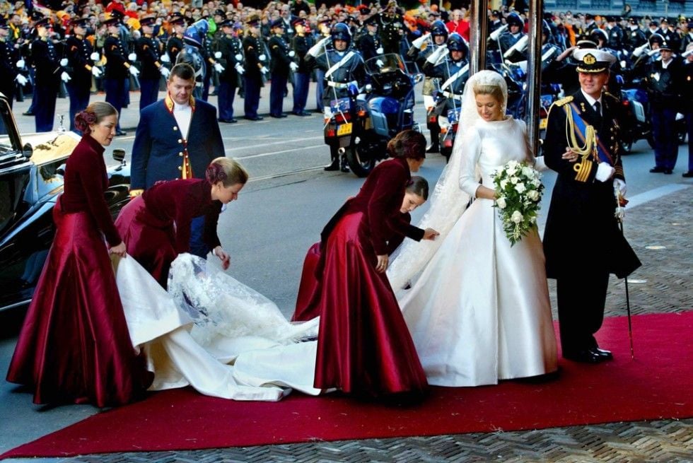 A bride in a white gown stands on a red carpet, surrounded by attendants in matching burgundy dresses adjusting her train. A groom in a decorated military uniform stands beside her; uniformed officers and a crowd are in the background.