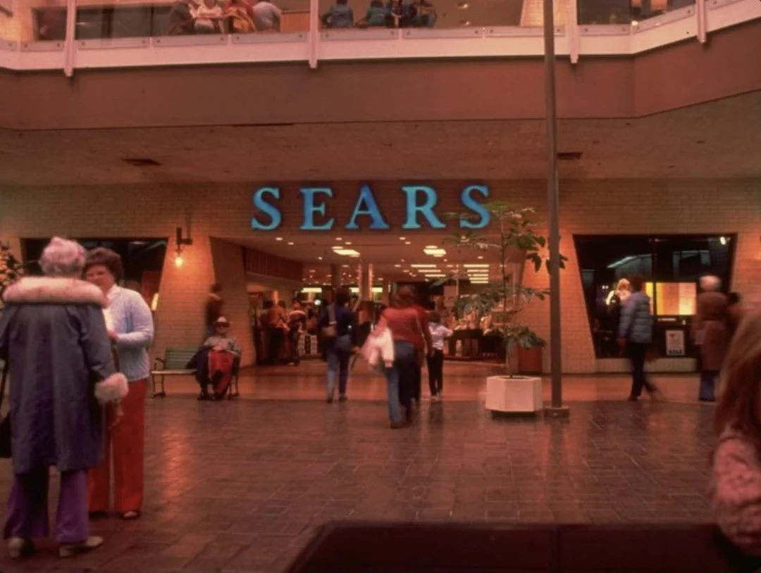People walk in front of a Sears store in a shopping mall. The large blue "SEARS" sign is prominent above the entrance, and various shoppers are visible, including an elderly woman in a blue coat and others in casual 1970s attire.