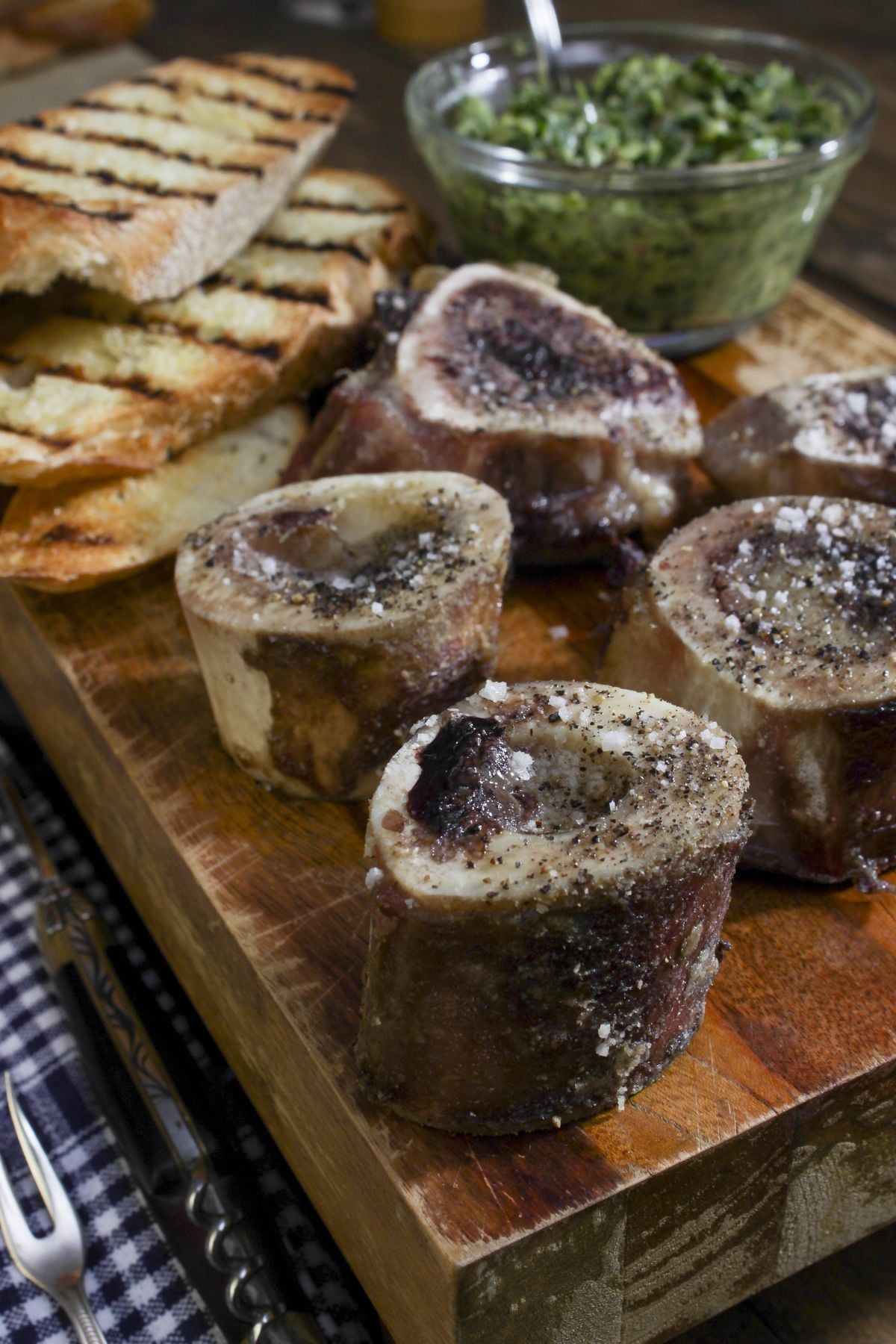 Four roasted bone marrow pieces topped with salt and pepper are arranged on a wooden board, accompanied by grilled bread slices and a bowl of green herb sauce in the background. Utensils and a checkered napkin are nearby.