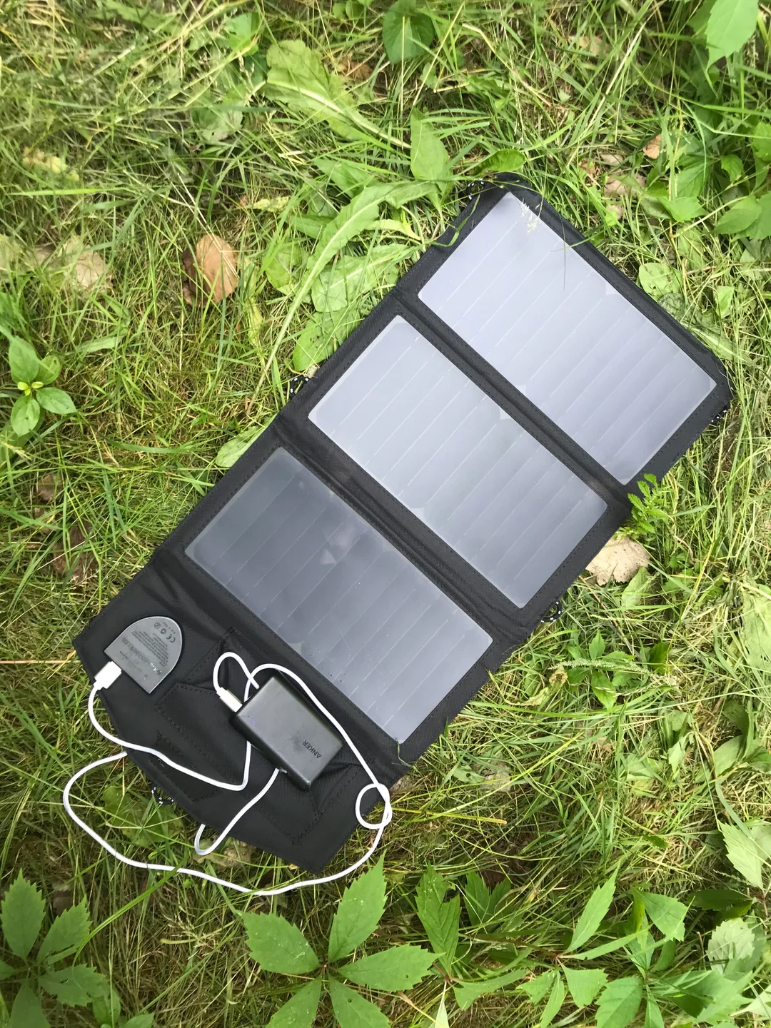 A portable solar panel charger with three panels and a connected device is lying on green grass and surrounded by leaves and plants outdoors.