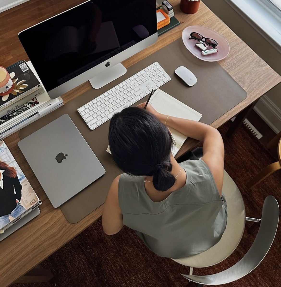 A person with dark hair in a low bun sits at a wooden desk, writing in a notebook. The desk has an iMac, MacBook, keyboard, mouse, magazines, glasses, keys, and a pink dish by a window.