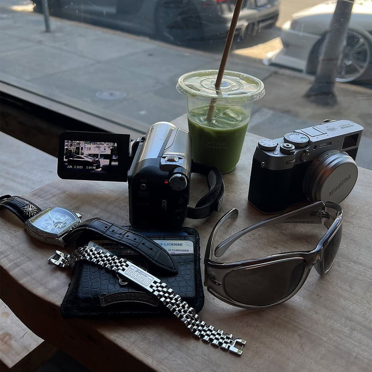 A wooden table with a camcorder, digital camera, green smoothie, sunglasses, wristwatch, wallet, license card, and a metal watch band, all placed by a bright window overlooking parked cars.