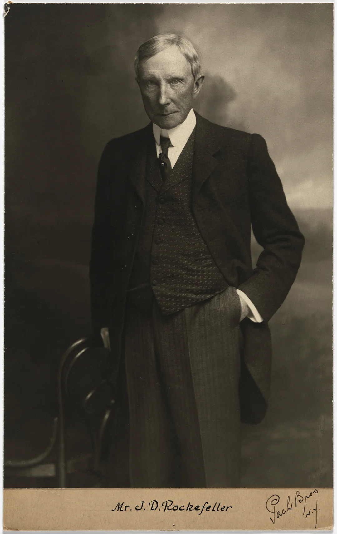 A stern-faced older man in a dark three-piece suit stands with one hand in his pocket and the other resting on a chair. The photo is sepia-toned and labeled "Mr. J. D. Rockefeller" at the bottom.