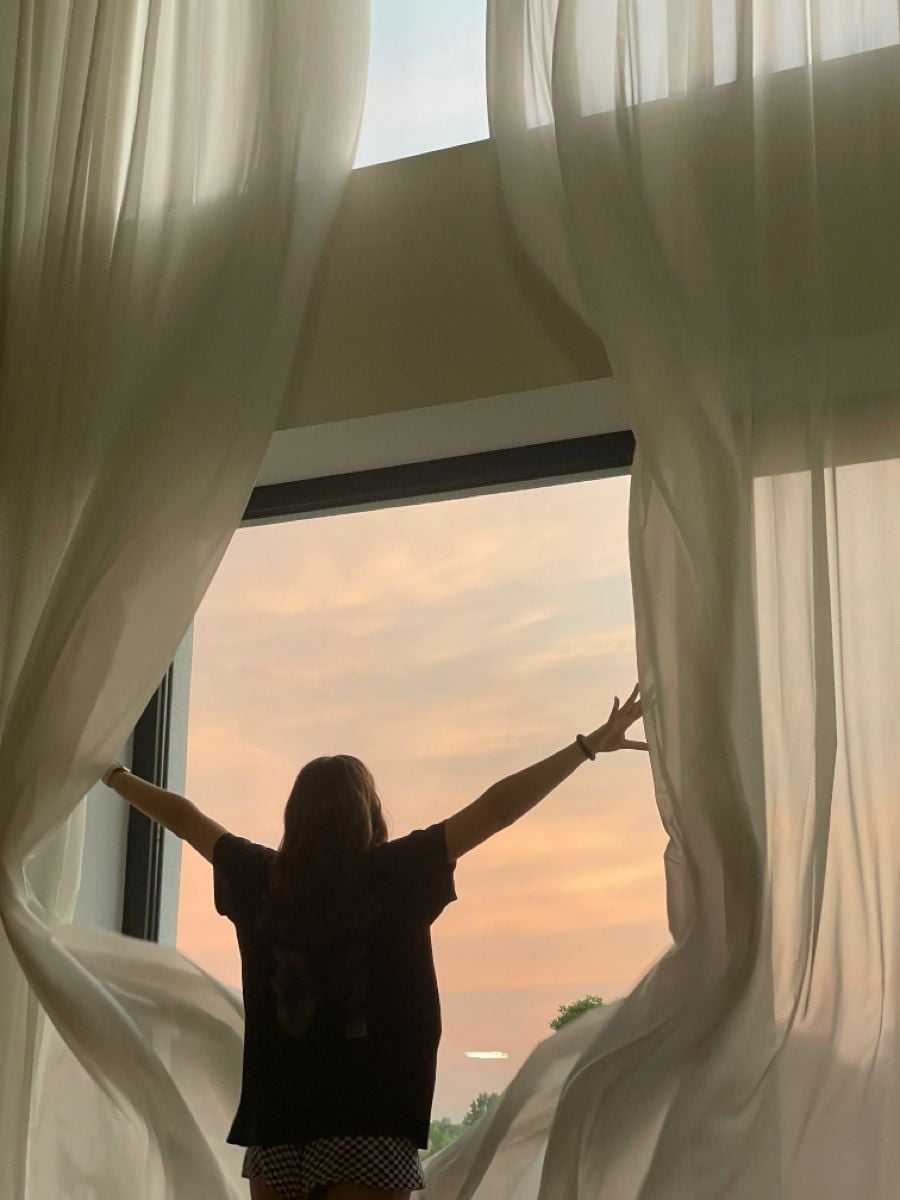 A person stands with arms outstretched, opening sheer curtains in front of a large window, looking out at a pastel-colored sunrise or sunset sky.
